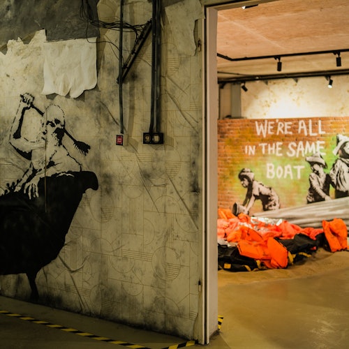 Espacio interior con mural de graffiti y una sala llena de chalecos salvavidas naranjas; el texto en la pared dice "TODOS ESTAMOS EN EL MISMO BARCO."