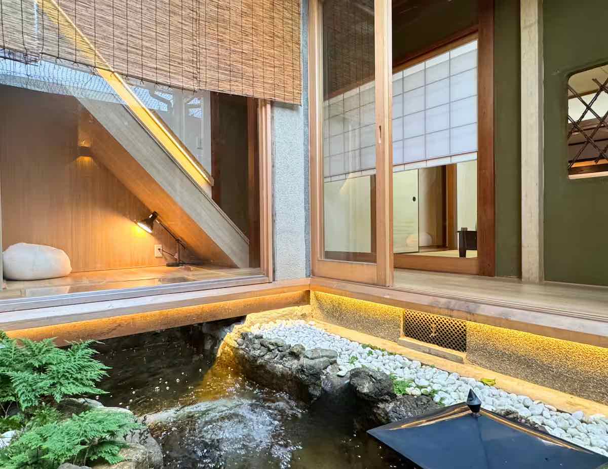 A tranquil Japanese-style indoor garden features a small koi pond with cascading water, surrounded by smooth white pebbles, bamboo accents, and wooden sliding doors leading to a serene tatami room.