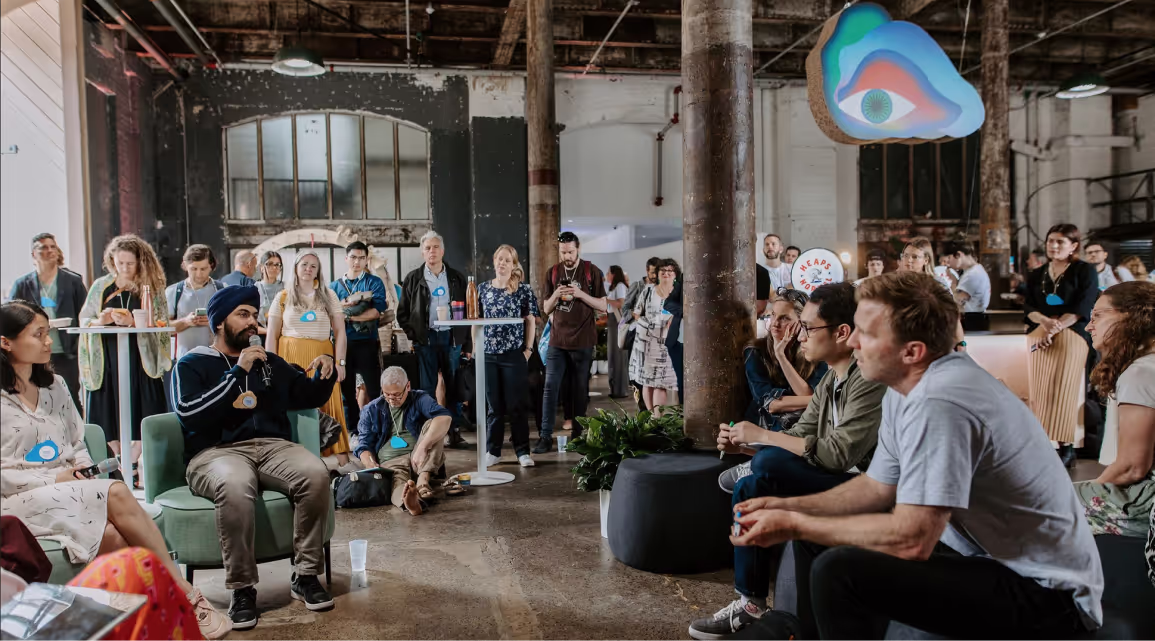 An audience seated on couches and floor cushions in a warehouse venue watching a talk, with a colourful hanging sculpture overhead