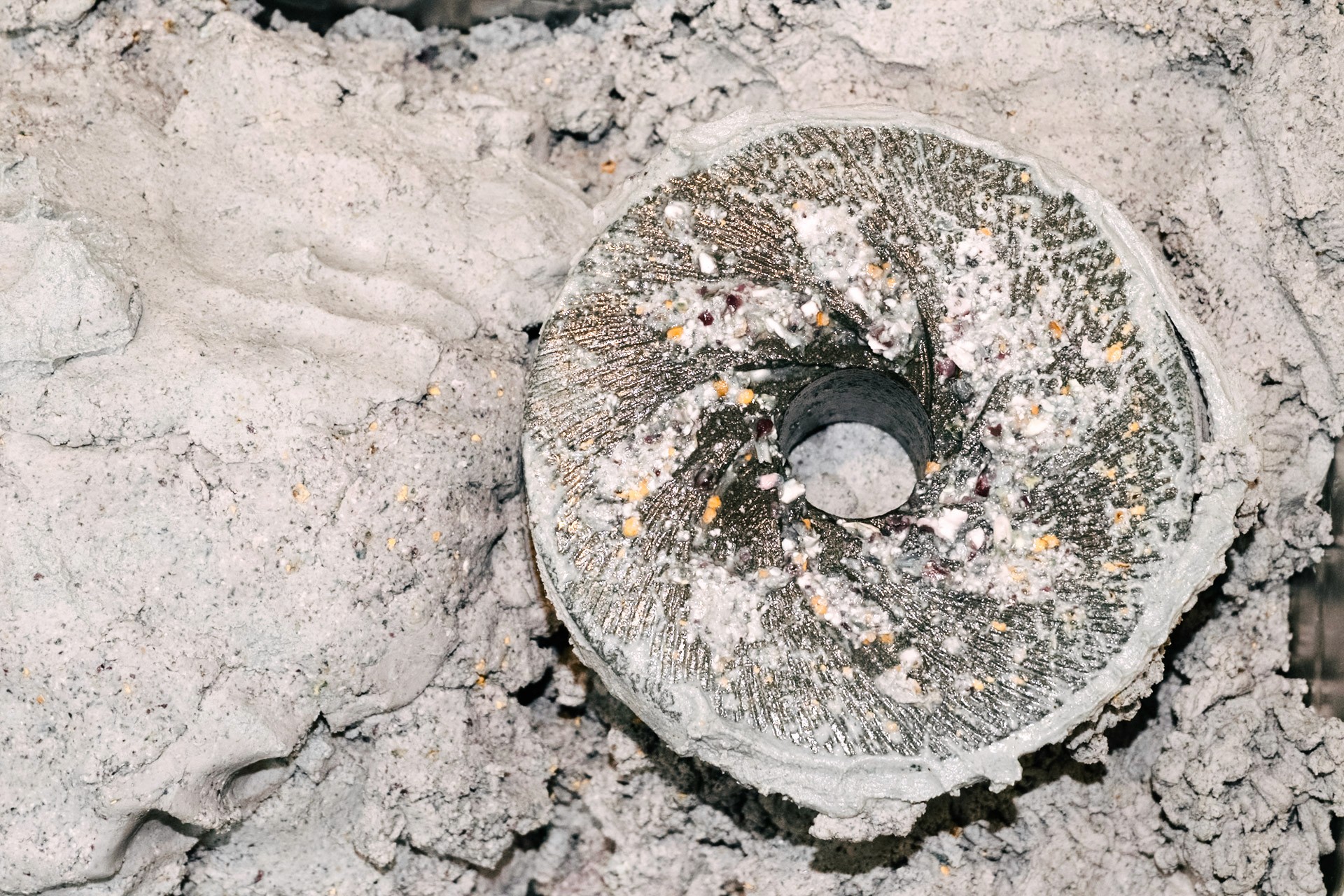 A round grinding stone resting face up in fresh grey masa, its cut grooves worn smooth in places.