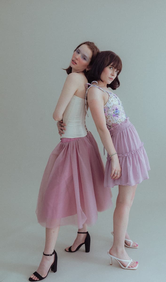 Models leaning away from each other in matching pink skirts, captured in a soft, creative pose during a natural light photoshoot at Revelator Studio in Shreveport.