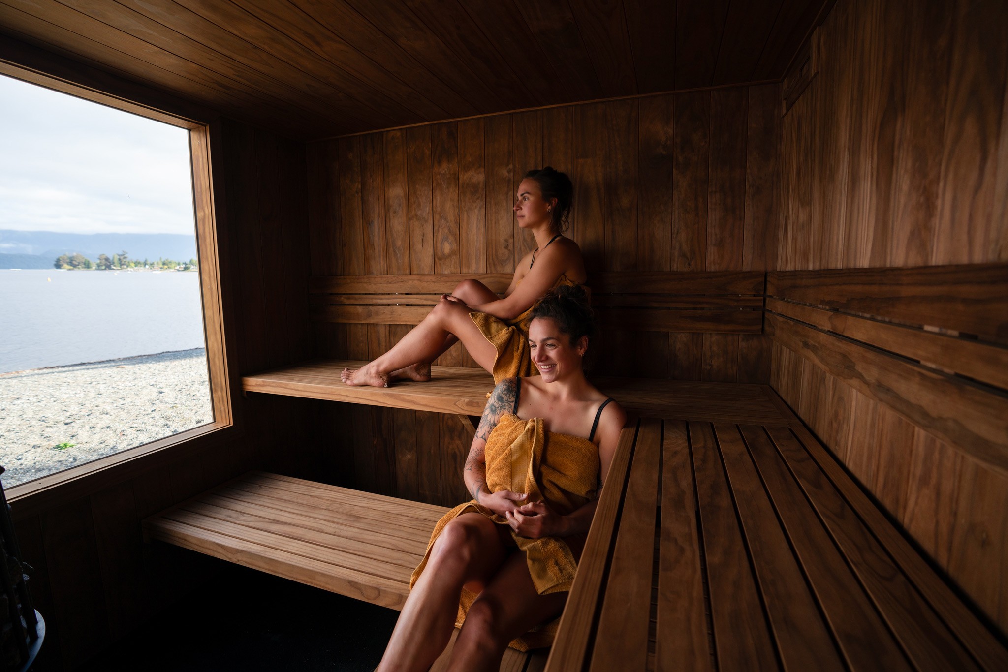 View from inside a cozy sauna with wooden benches and a heater in the foreground, overlooking a tranquil lake and misty mountains in the background.