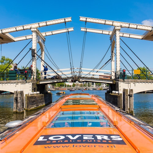 Orange canal boat with "LOVERS" logo approaching a white drawbridge over a canal, under a clear blue sky.