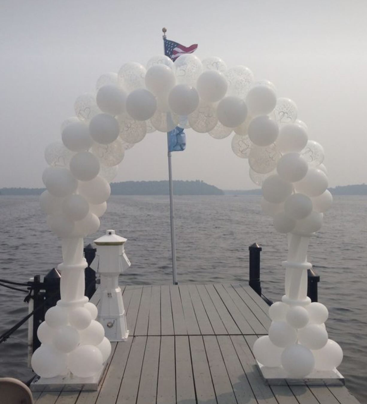 A white balloon arch stands prominently on a wooden dock extending over a tranquil lake, with a flagpole displaying an American flag in the background against a hazy sky.