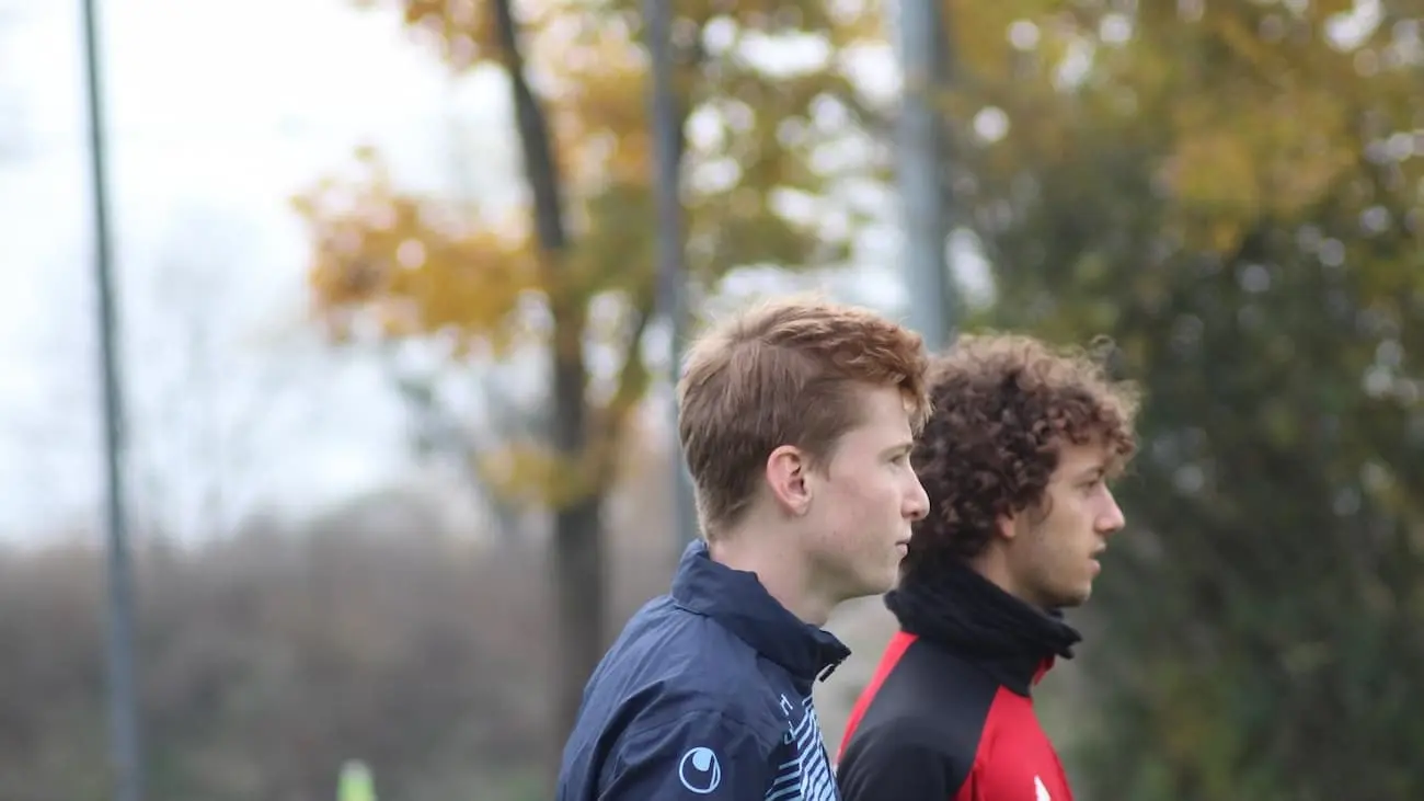 Two football coaches watch the game closely and discuss tactical decisions that influence the development of their players and the team's performance. Autumnal trees can be seen in the background, placing the picture in a season and emphasising the focus on the game.