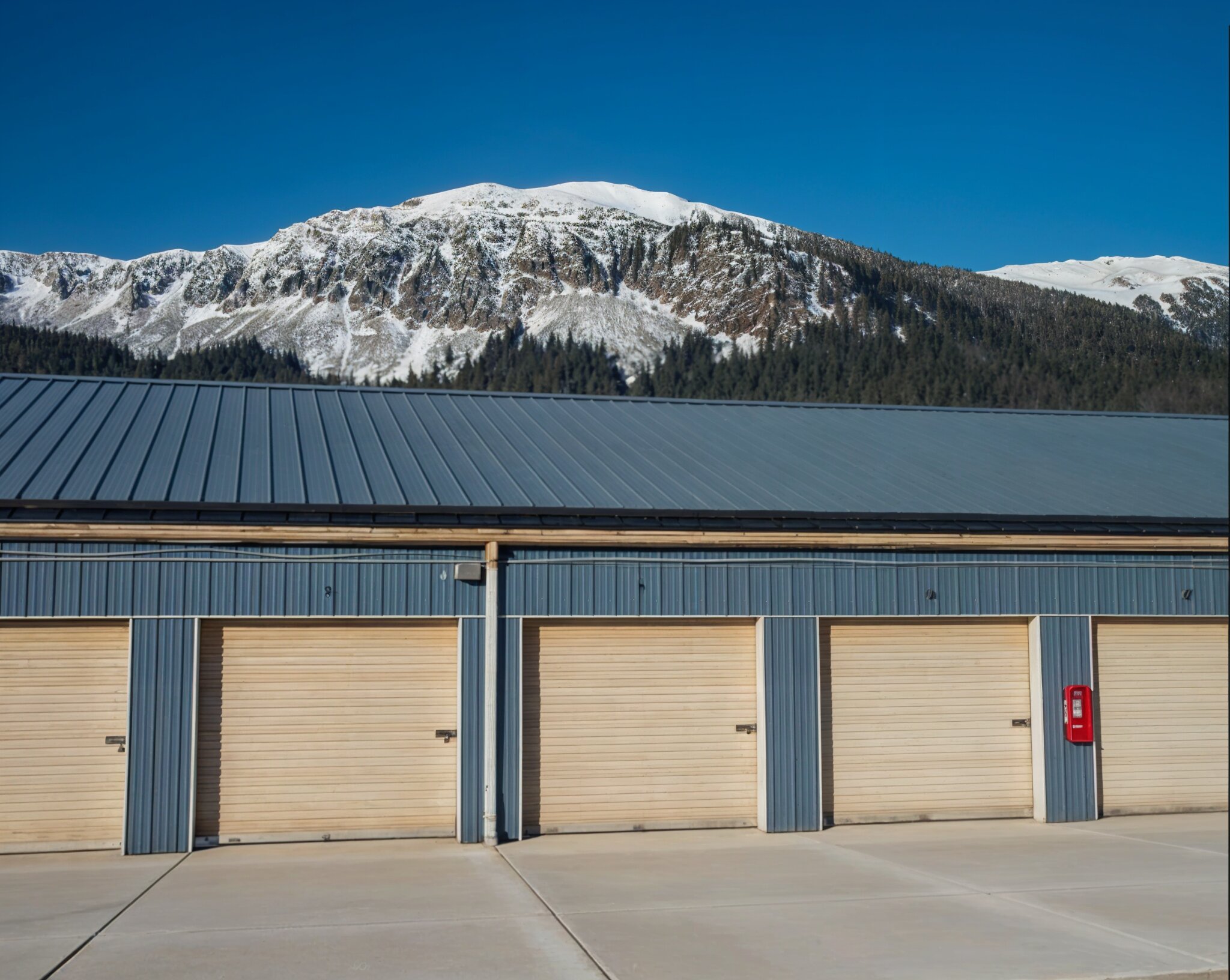 Aerial view of a self-storage facility with a two-story grey board-and-batten office building in the foreground, a surface parking lot with accessible striping, and a row of single-story storage units with blue roll-up doors extending along the left under a partly cloudy sky.