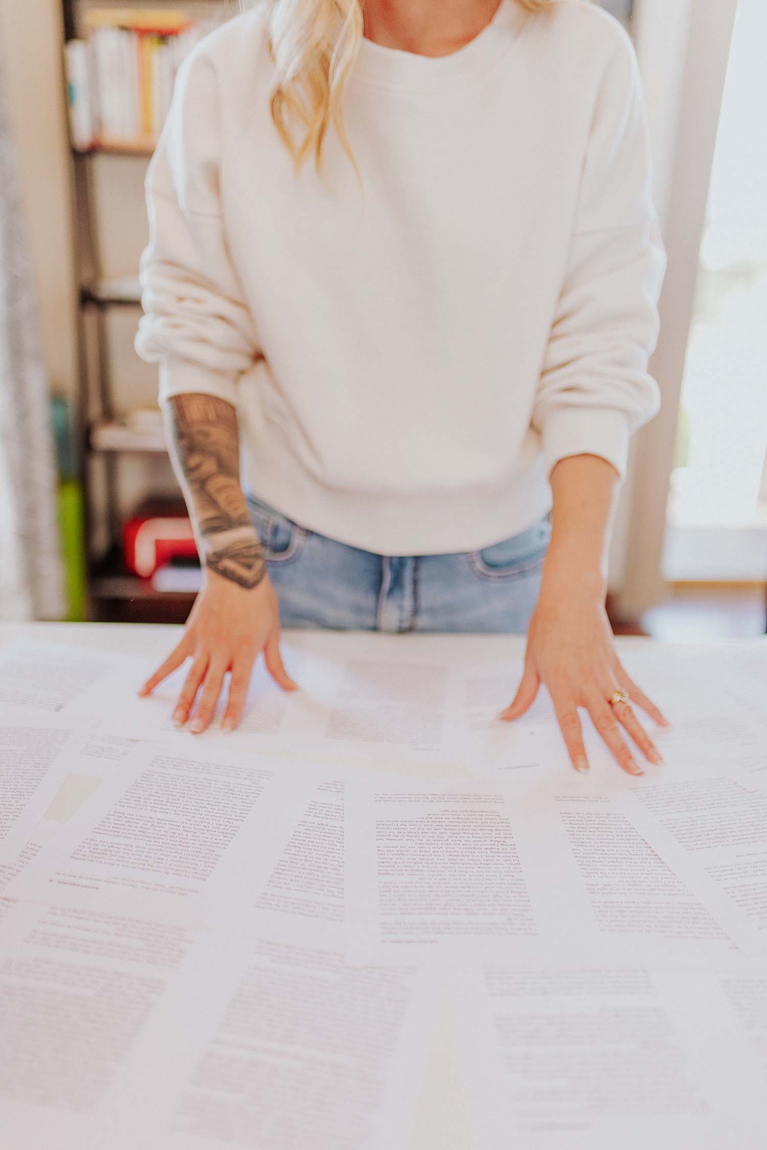 A view downwards of a woman's torso standing in front of a table with many pages of a book manuscript spread out on it, her hands are spread over the pages