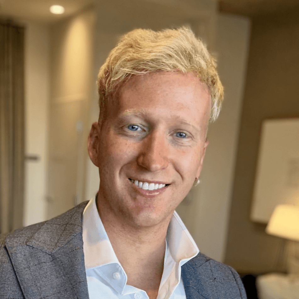 A young man with light brown hair is wearing a black suit jacket and white shirt, standing in a softly lit office setting, conveying professionalism.