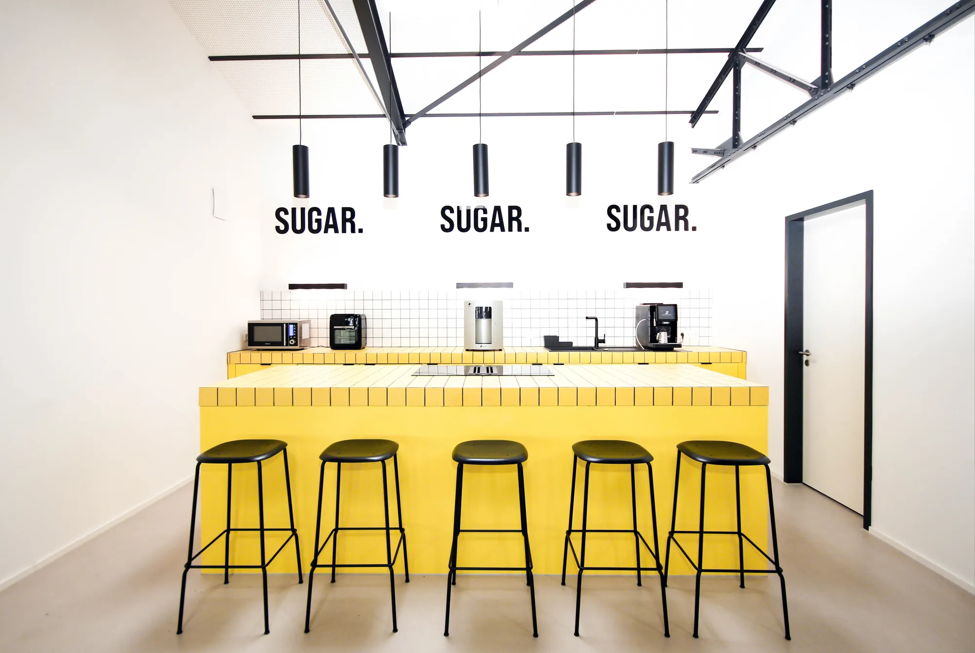 Yellow tiled kitchen counter with bar stools under SUGAR lettering in the bright loft.