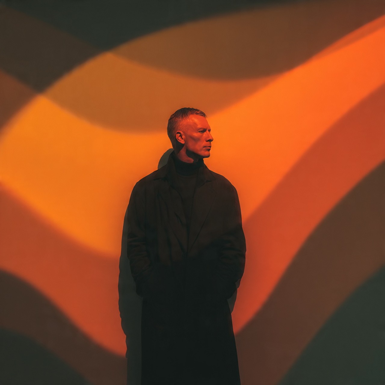A person stands against a vibrant background of orange and yellow wavy patterns. They are wearing a dark coat, looking contemplative and thoughtful.