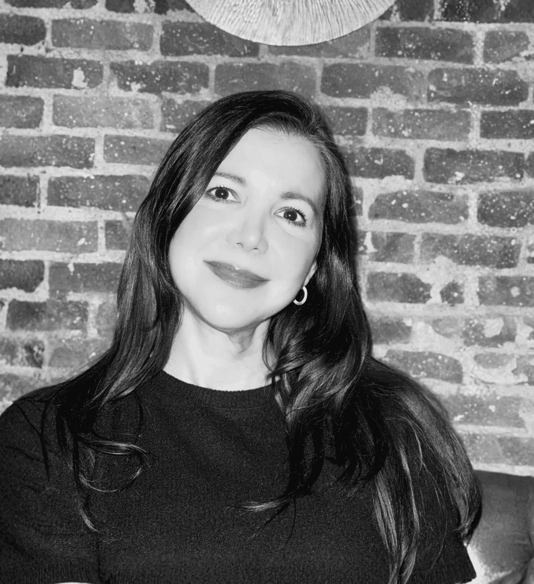 A smiling woman with long dark hair & dark sweater poses against a brick wall.