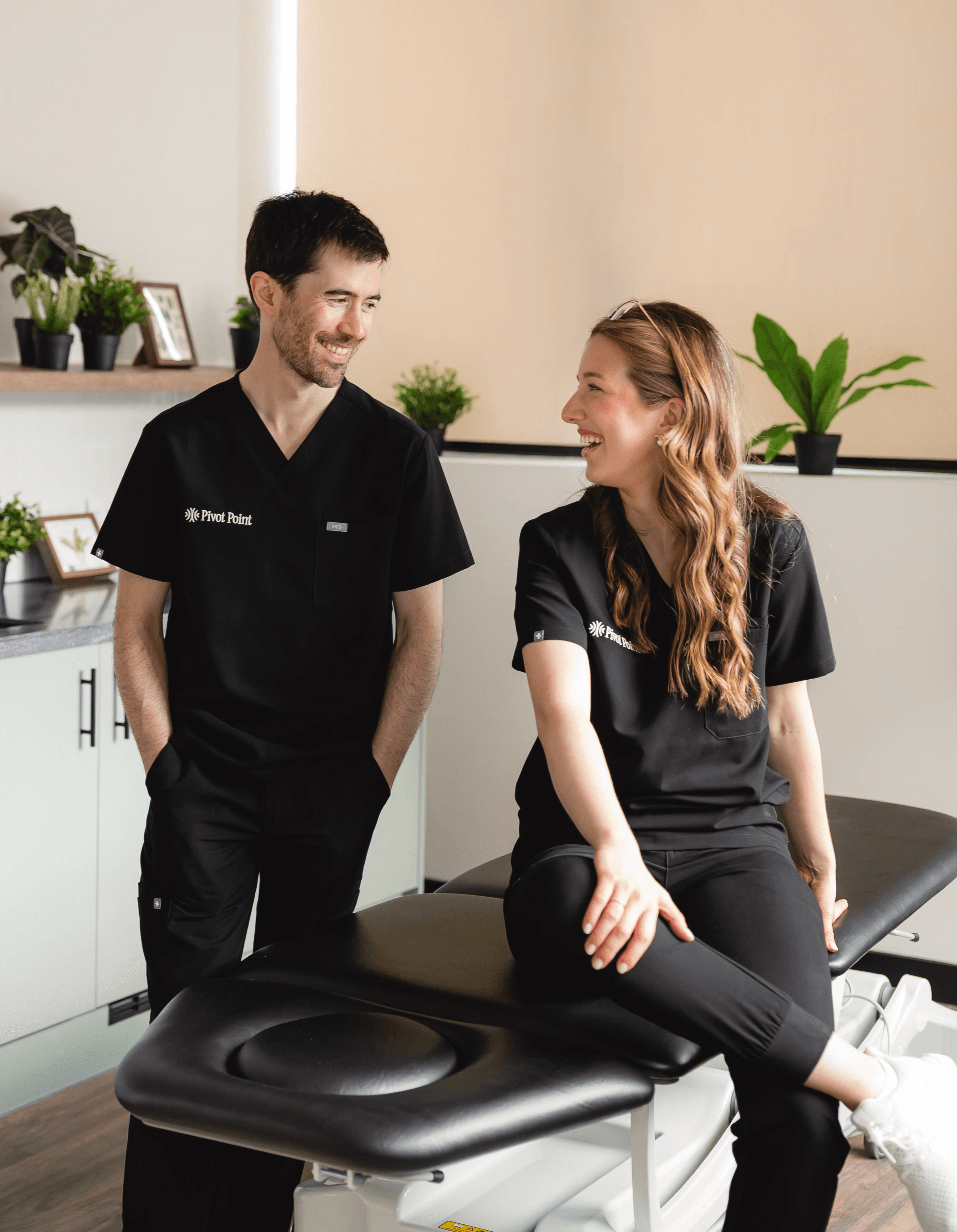 Two physiotherapists in uniform in a treatment room, facing each other and smiling