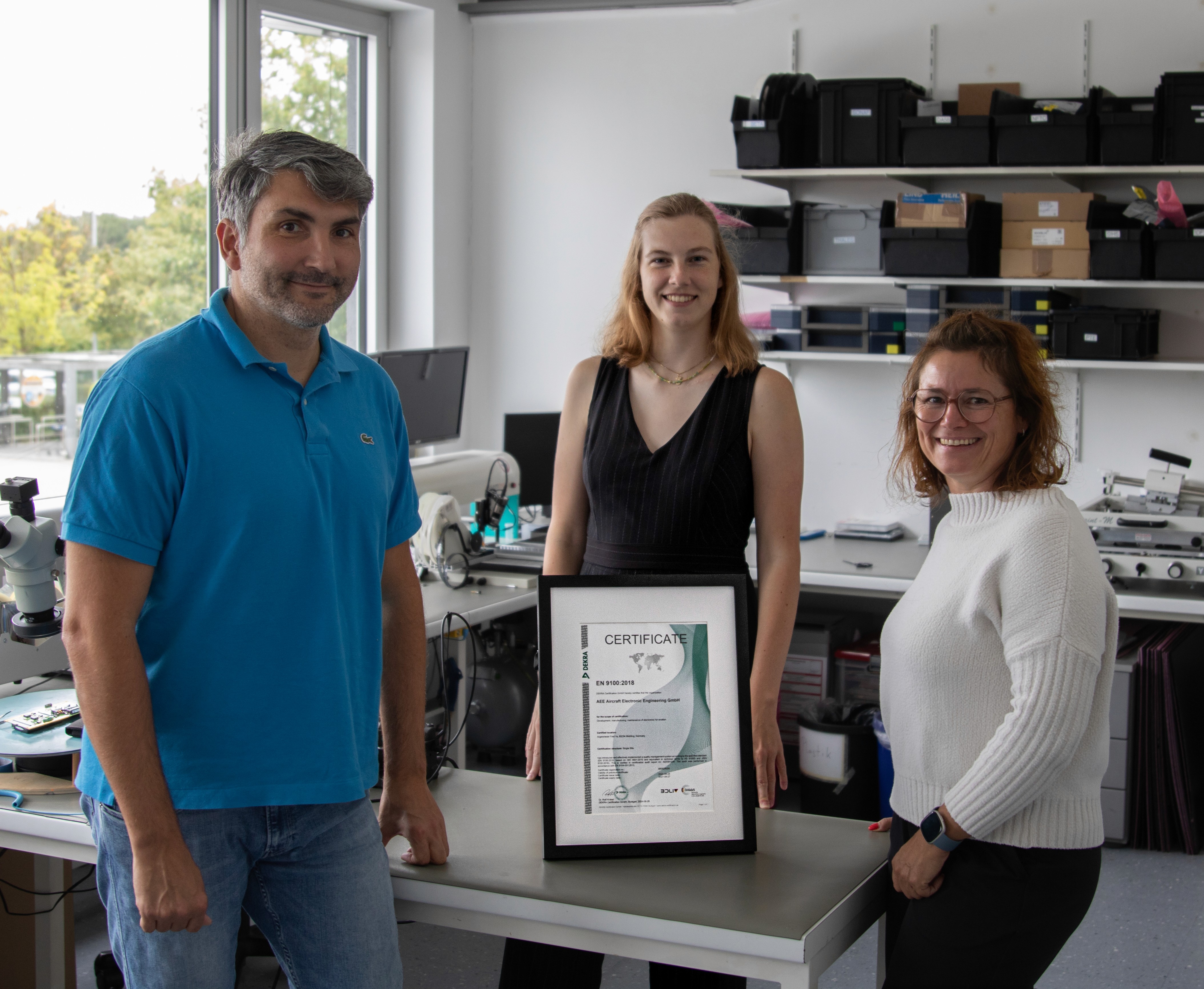 Two women and one man in a laboratory, standing around a table that holds a framed certificate.