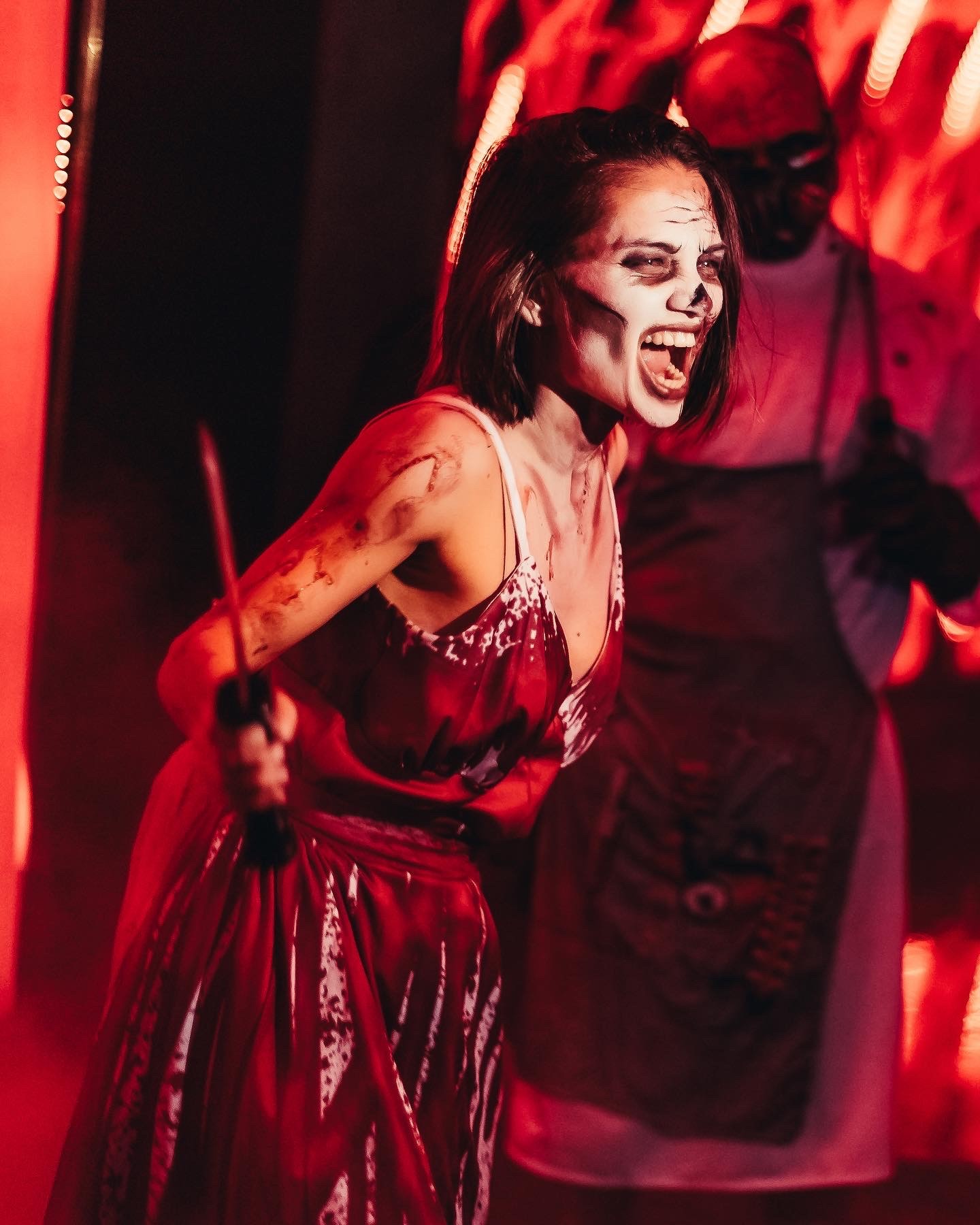 Halloween scare actor in bloody red dress holding knife prop with masked horror character at Stage nightclub Dubai