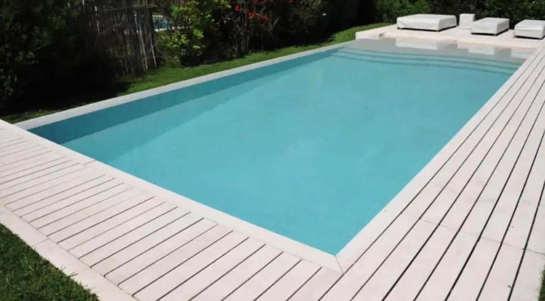 Moderna pileta rectangular rodeada de un elegante deck de madera pintado de blanco, con zona de solárium y reposeras minimalistas.