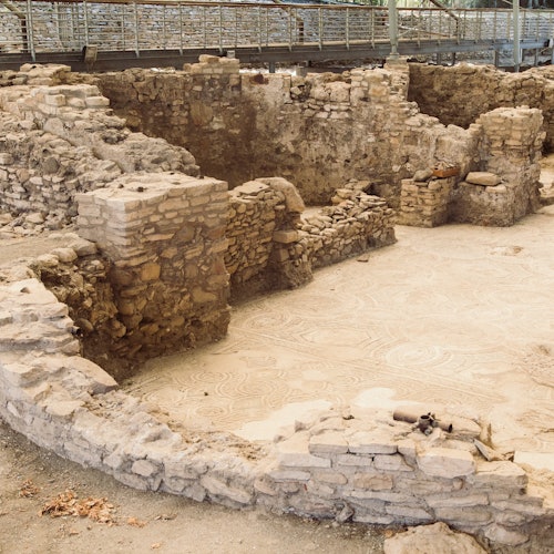古老的石头遗址，部分墙壁仍然存在，且有可见的马赛克地板。背景中有一条高架人行道。