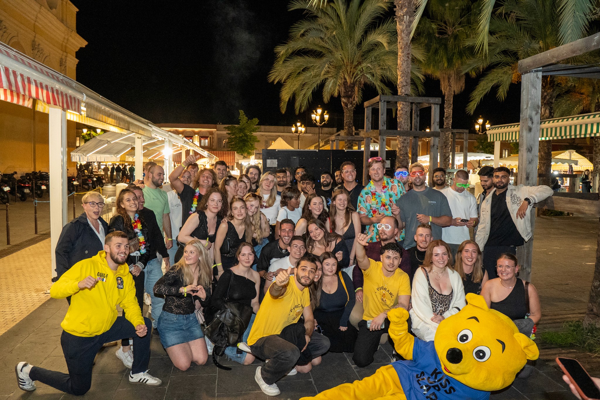 Large international group of young travelers posing together outdoors at night during a bar crawl in Nice surrounded by palm trees and city lights showcasing a fun social atmosphere group bonding and the vibrant nightlife experience of the French Riviera