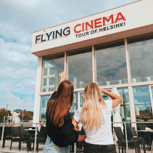 Kaksi pitkähiuksista naista seisoo rakennuksen edessä, jossa lukee "Helsingin lentävä elokuvakierros,” ja he katsovat ylös ja osoittavat kylttiä.