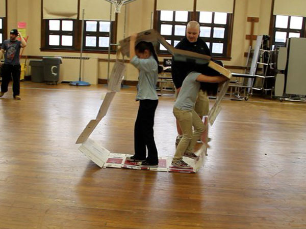 Three individuals are in a gymnasium with light brown wooden flooring. Two are positioned inside a large cardboard structure resembling a house or building frame. One person stands inside the structure, holding onto the frame, while another stands on the frame’s base. A third person stands to the left of the structure, observing. The gymnasium has large windows and exercise equipment visible in the background.