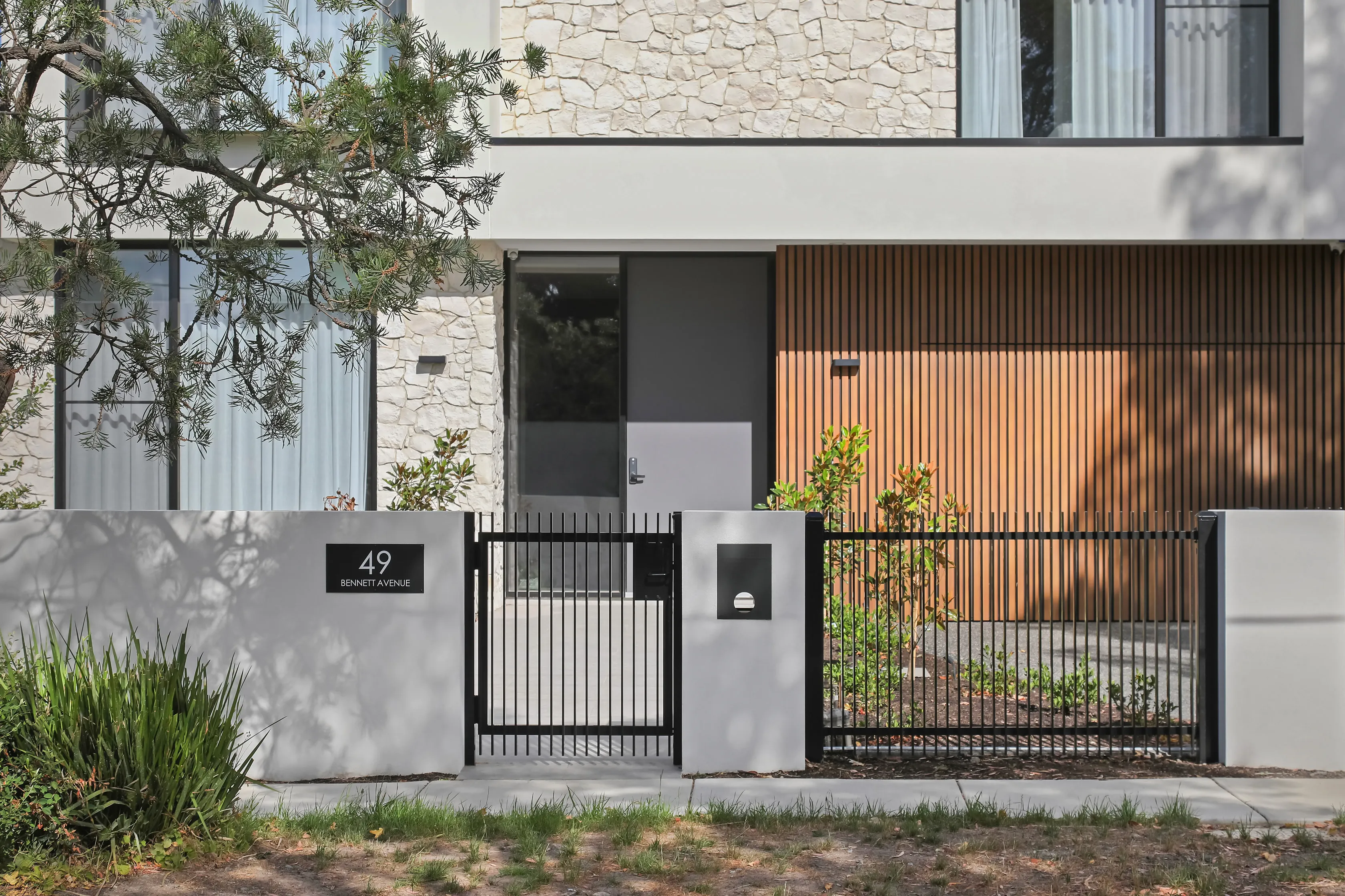 Front entrance of Mount Waverley Residence with stone cladding, timber-batten wall, dark pivot door and the number 49 Bennett Avenue sign
