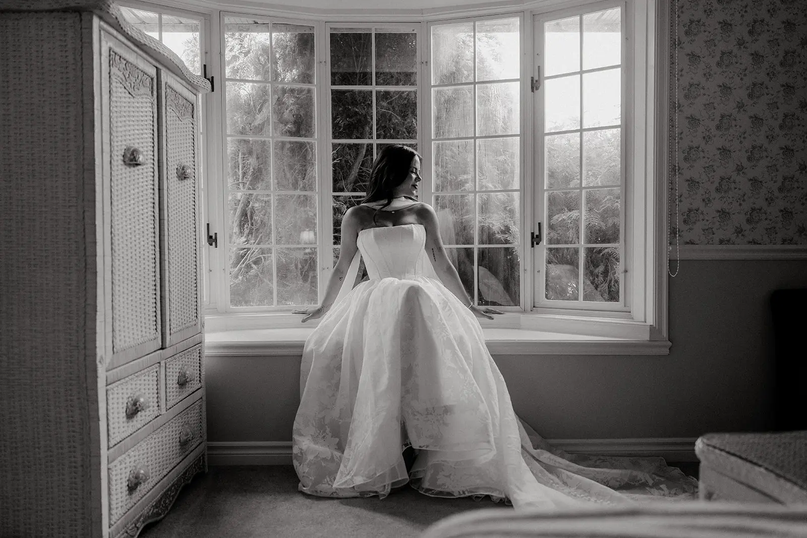 Black-and-white portrait of a bride seated in a window alcove wearing a strapless wedding gown.