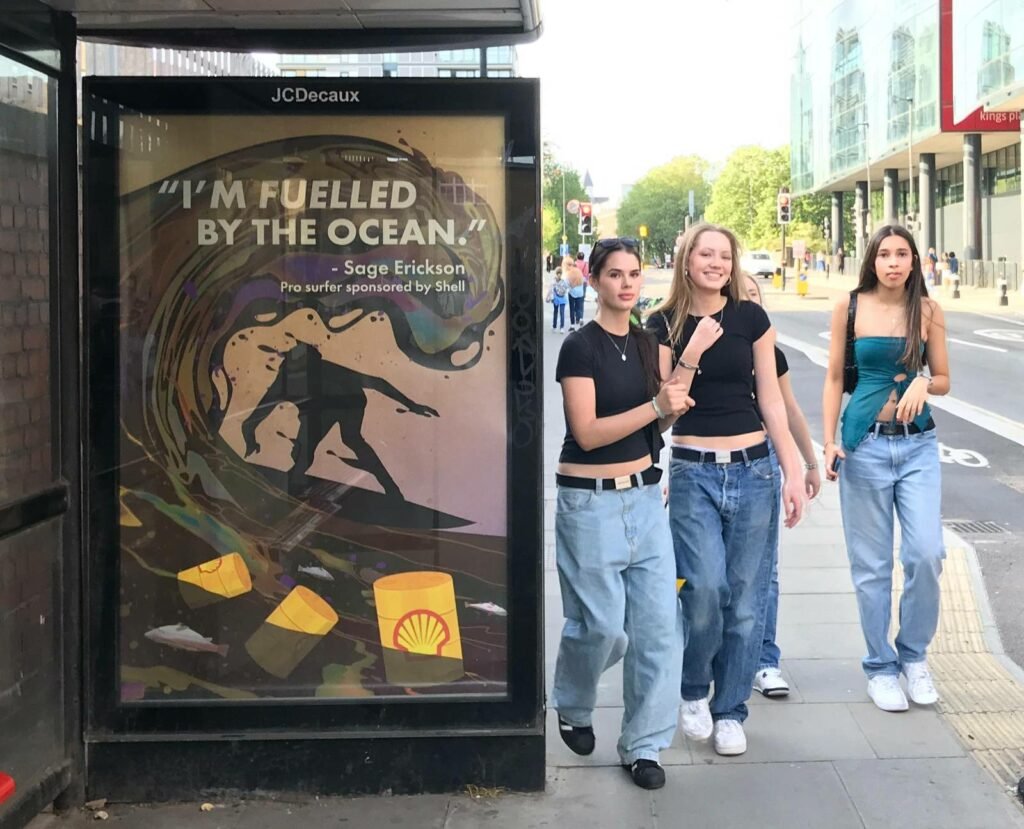 photo of 3 teenagers next to a bus stop Poster showing an illustration of a surfer covered in fuel, next to the words 'I'm fuelled by the Ocean'. On the water are floating barils of oil.