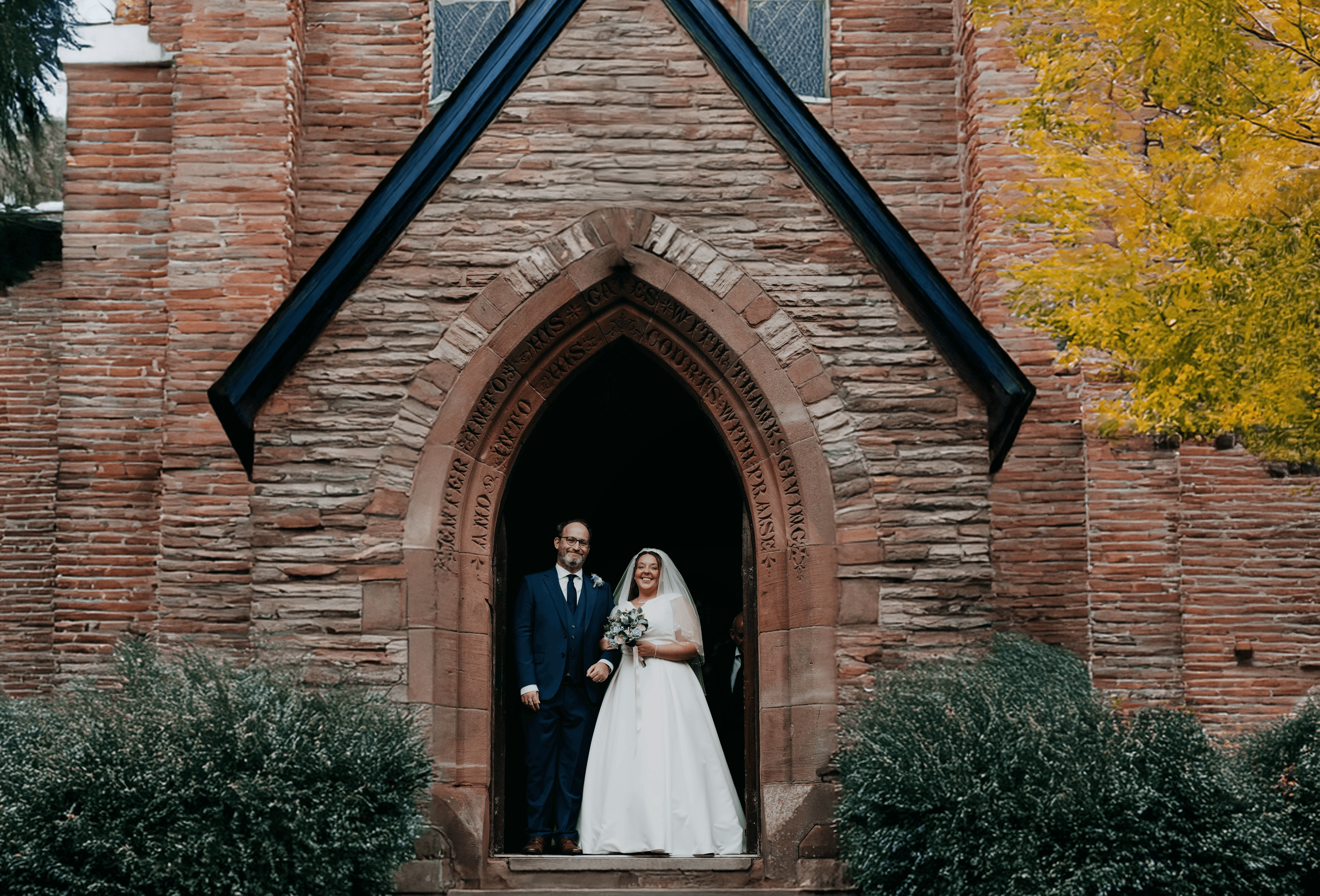 The bride and groom stand together, smiling, framed perfectly within the arched stone doorway of the church just after their ceremony.