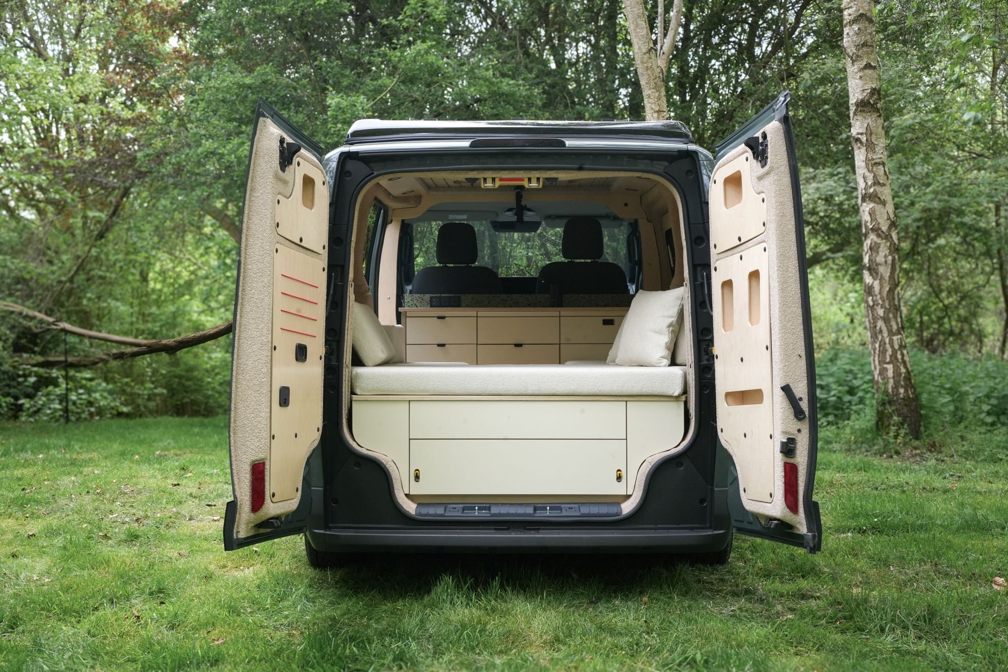 The back of a camper van with open doors, showing a living space with a bed and storage drawers, parked on green grass in a wooded area.
