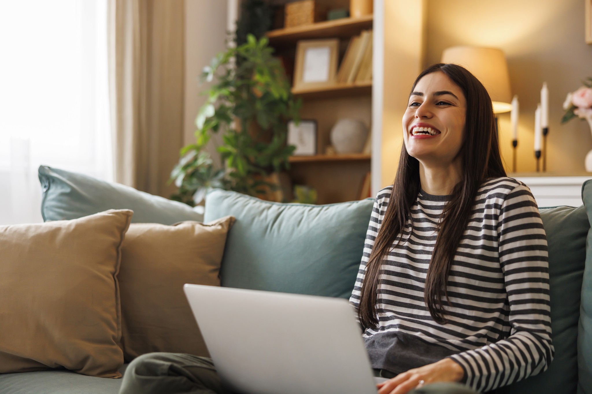 Happy Young Woman Using Laptop While Relaxing on Sofa at Home