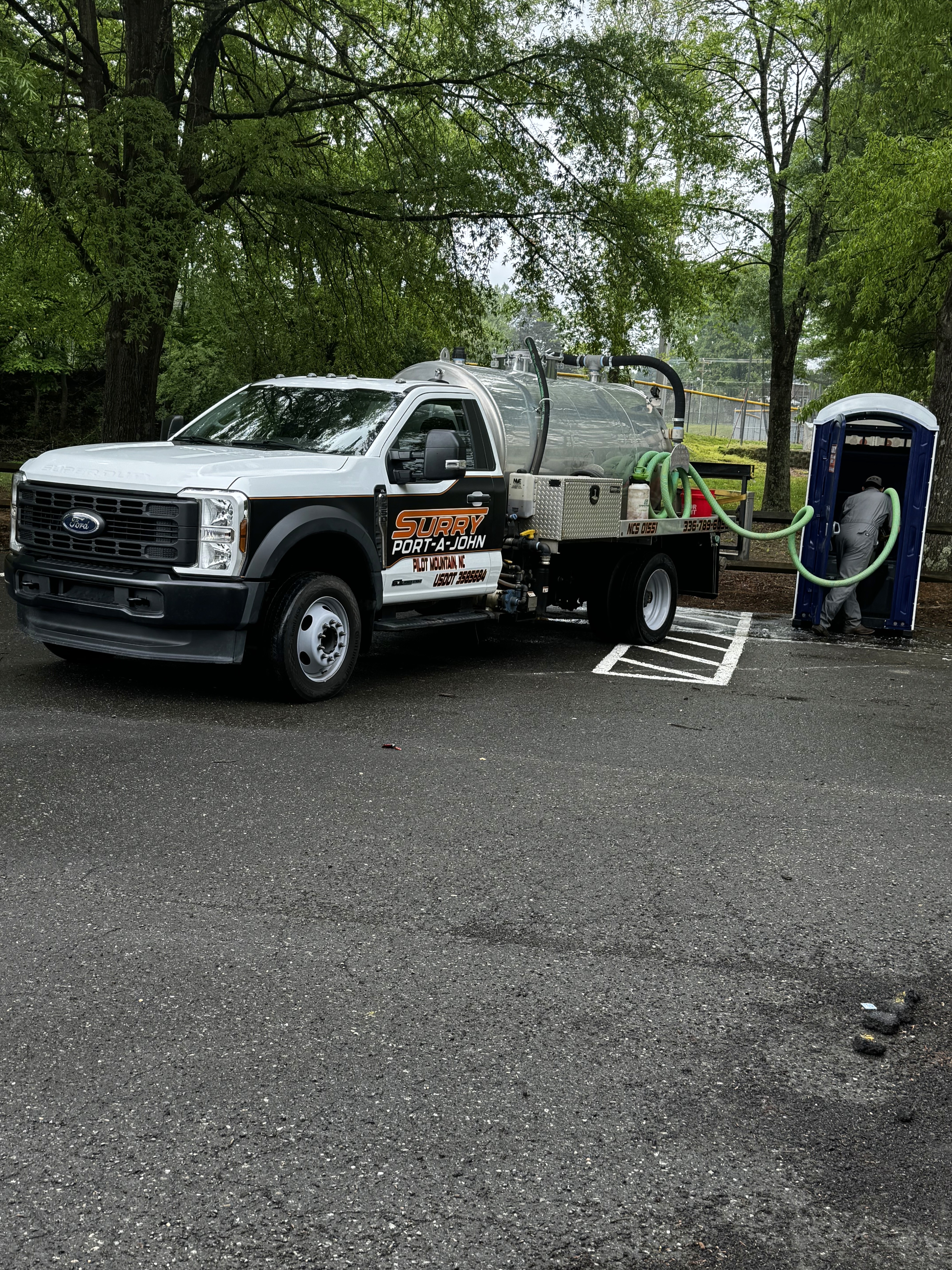 A white utility truck labeled "Surry Port A John Rental" is parked on a wet, paved surface near a portable toilet among green trees, with hoses connected for service.