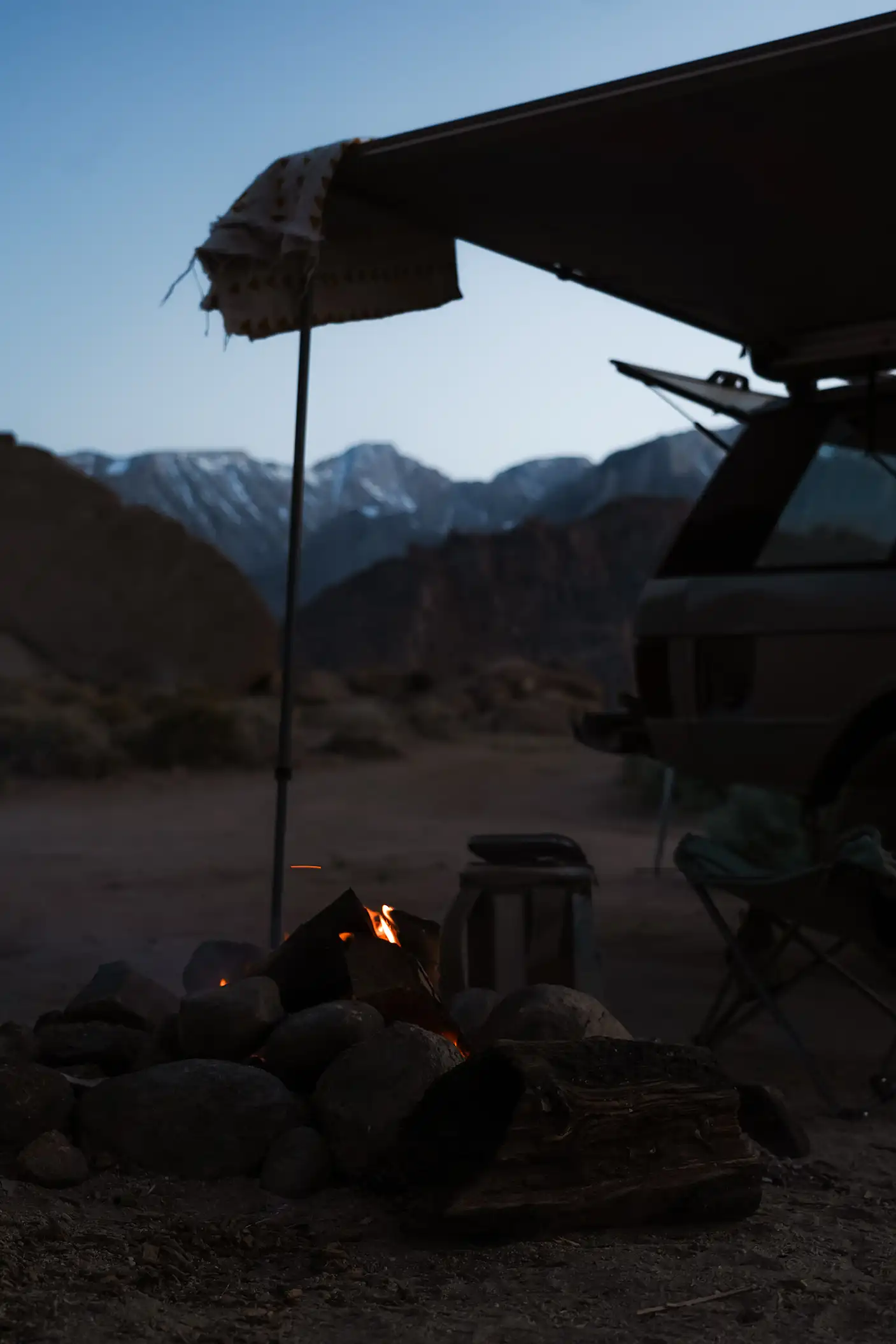 A silver Range Rover SUV parked at a campsite, with a campfire glowing at dusk.