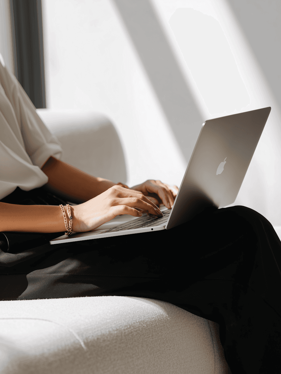 A person is typing on a laptop while sitting on a couch, with sunlight casting shadows.