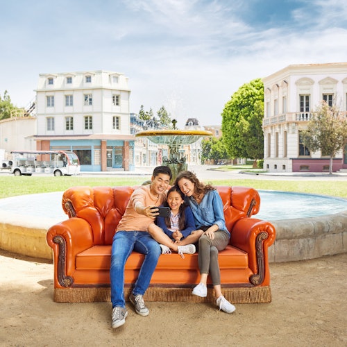 A family of three sits on an orange couch outdoors, taking a selfie. Buildings and trees are visible in the background.