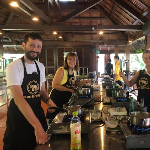 Tiga orang berada di dapur berbentuk desa, memakai apron, memasak di stesen individu di bawah siling kayu yang tinggi dengan lampu gantung.