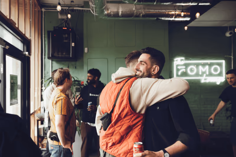 Two members hugging in the FOMO House bar, the FOMO neon behind them