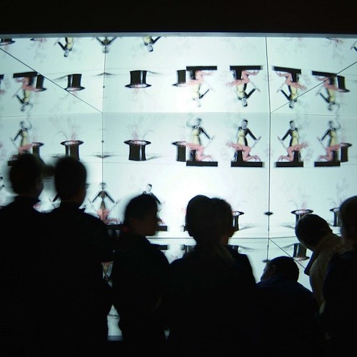 Silhouetted people stand in front of a large illuminated screen displaying repeated images of a performer with a top hat.