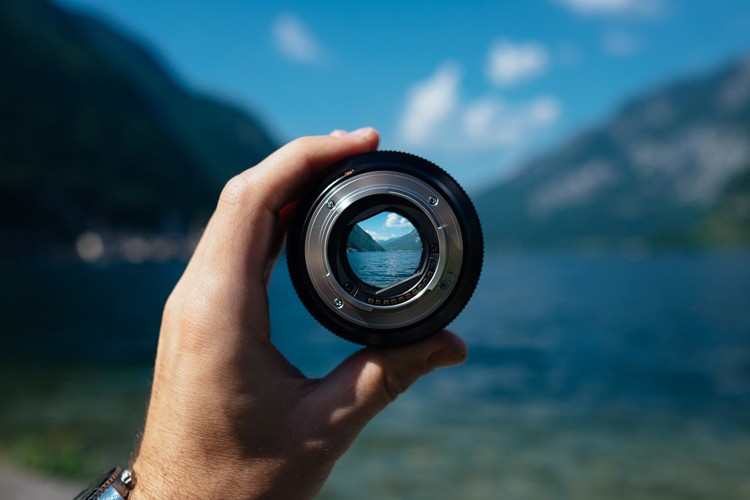A person holding a camera lens with a lake and mountains in the background