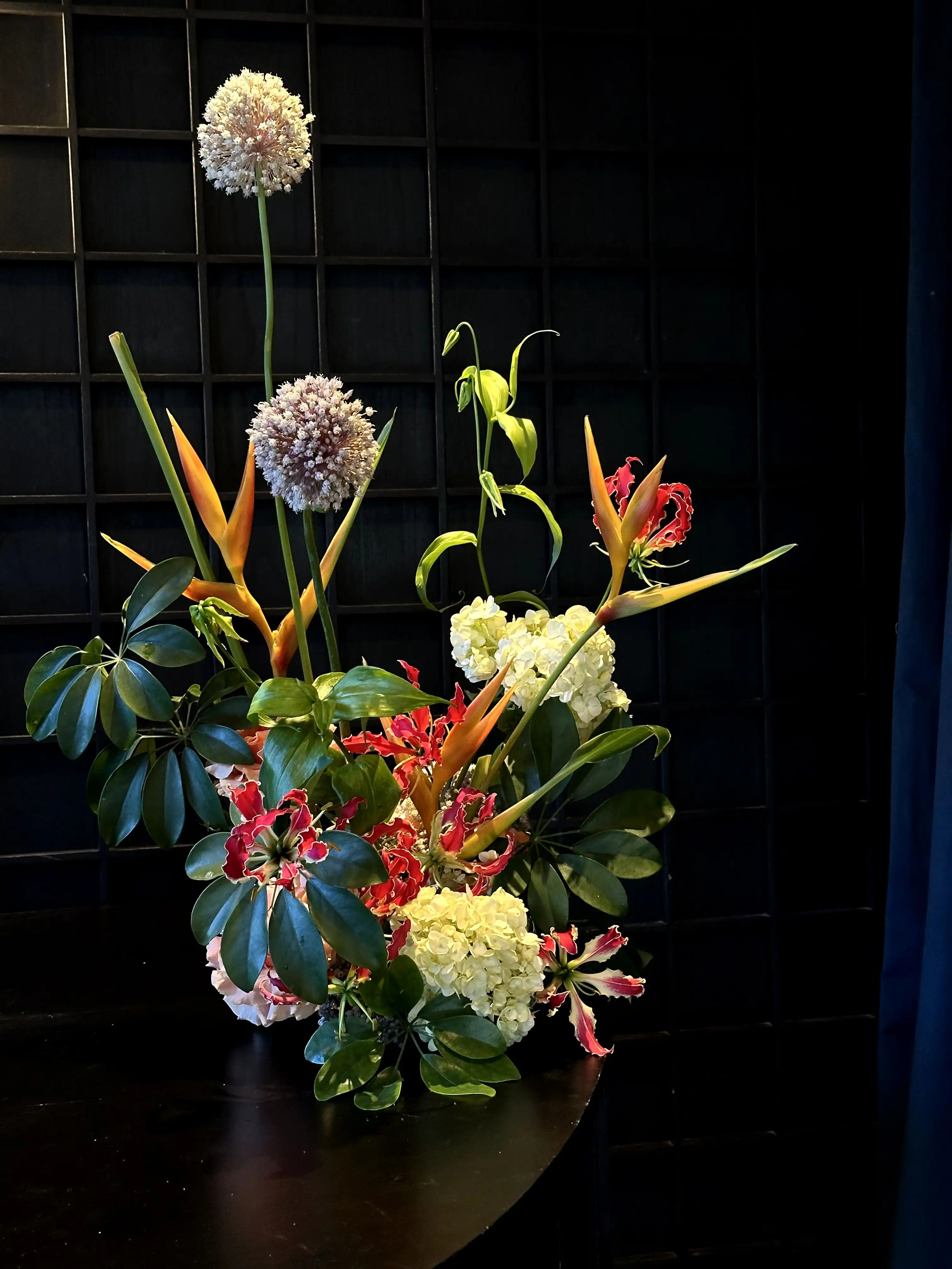 Arreglo floral contemporáneo con flores exóticas, incluyendo aves del paraíso, hortensias blancas, gloriosas rojas y flores esféricas lilas, dispuesto sobre una mesa negra frente a un fondo de paneles oscuros.