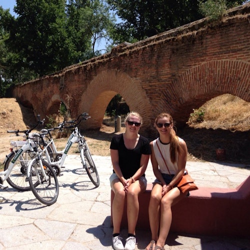 Üç bisikletin park ettiği, ağaçlarla çevrili, tuğla kemer şeklindeki bir köprünün önünde bir bankta oturan iki kişi.