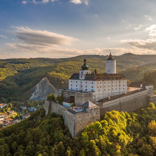 白色城墙和多座塔楼的山顶城堡，周围环绕着郁郁葱葱的森林，下方山谷中可以看到一个村庄。