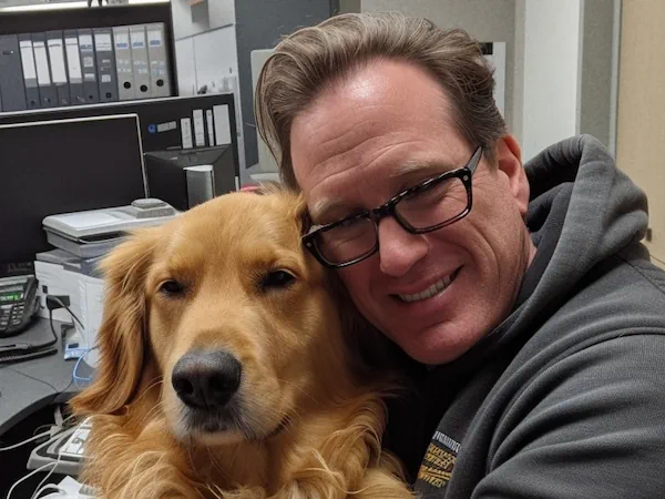 A man in a gray hoodie wearing glasses and hugging a dog