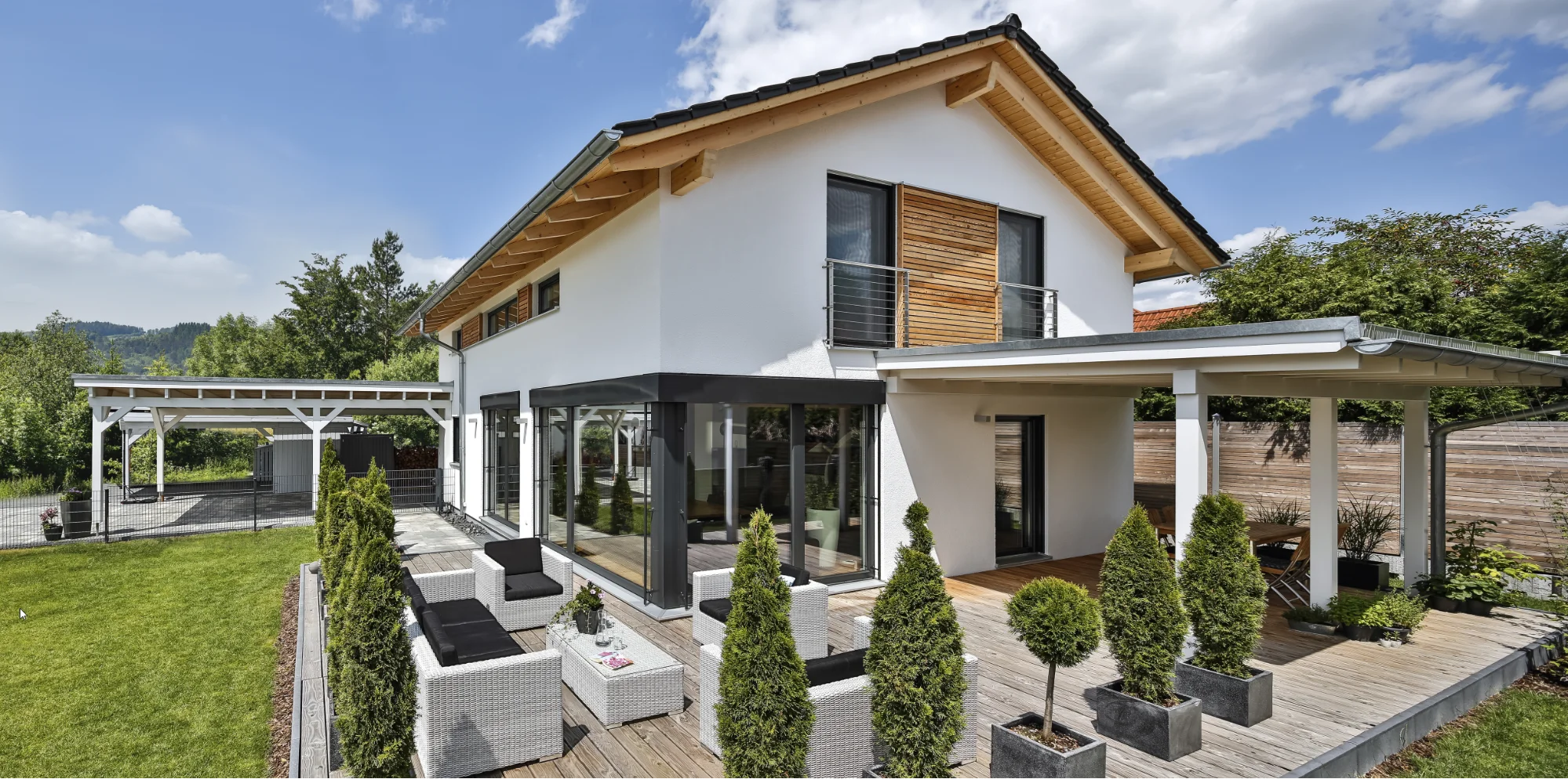 Modernes Familienhaus mit großer Holzterrasse, Poolhaus und Carport im Grünen.
