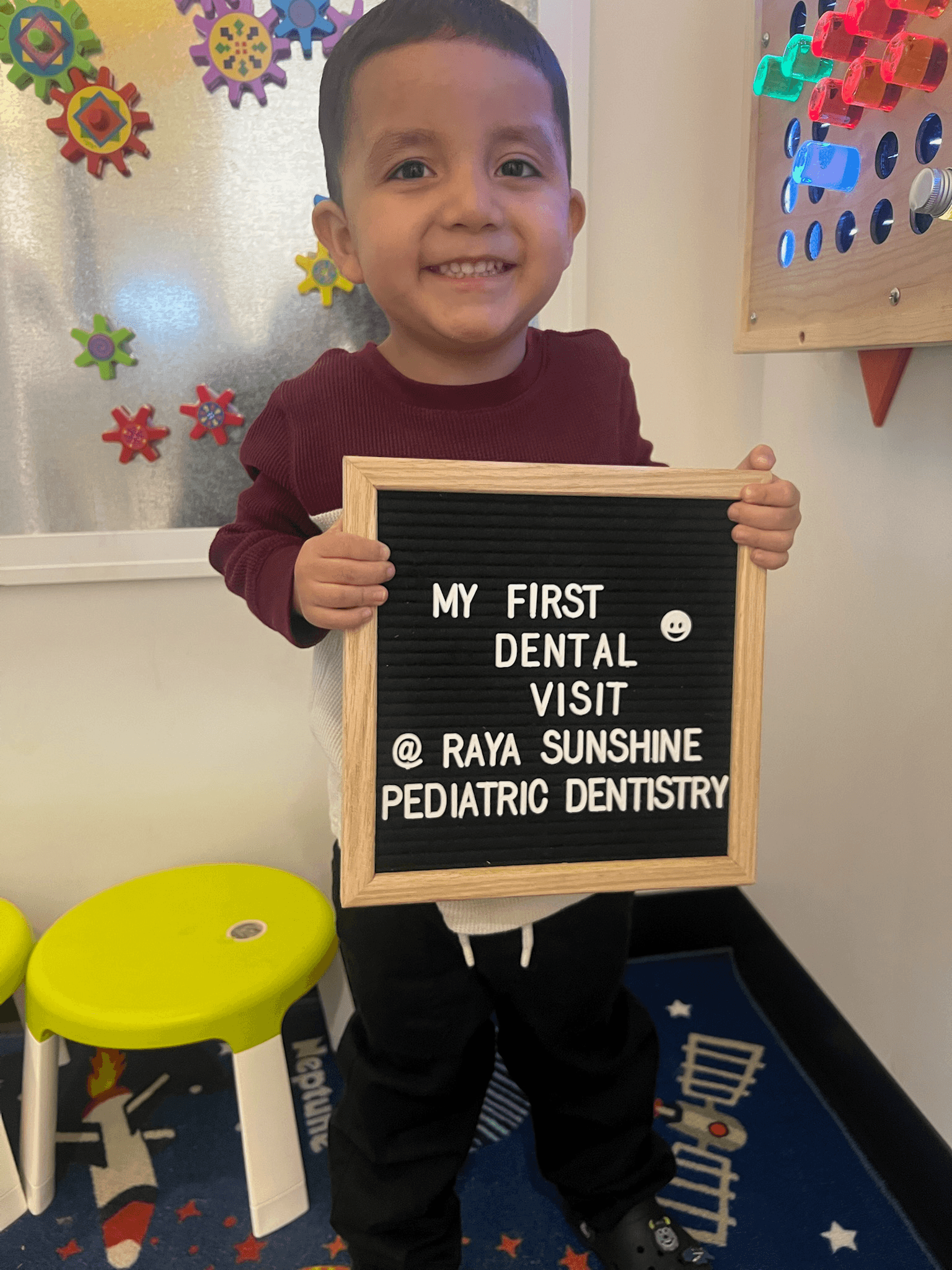 A smiling child standing indoors holding a sign that reads “My first dental visit at Raya Sunshine Pediatric Dentistry."