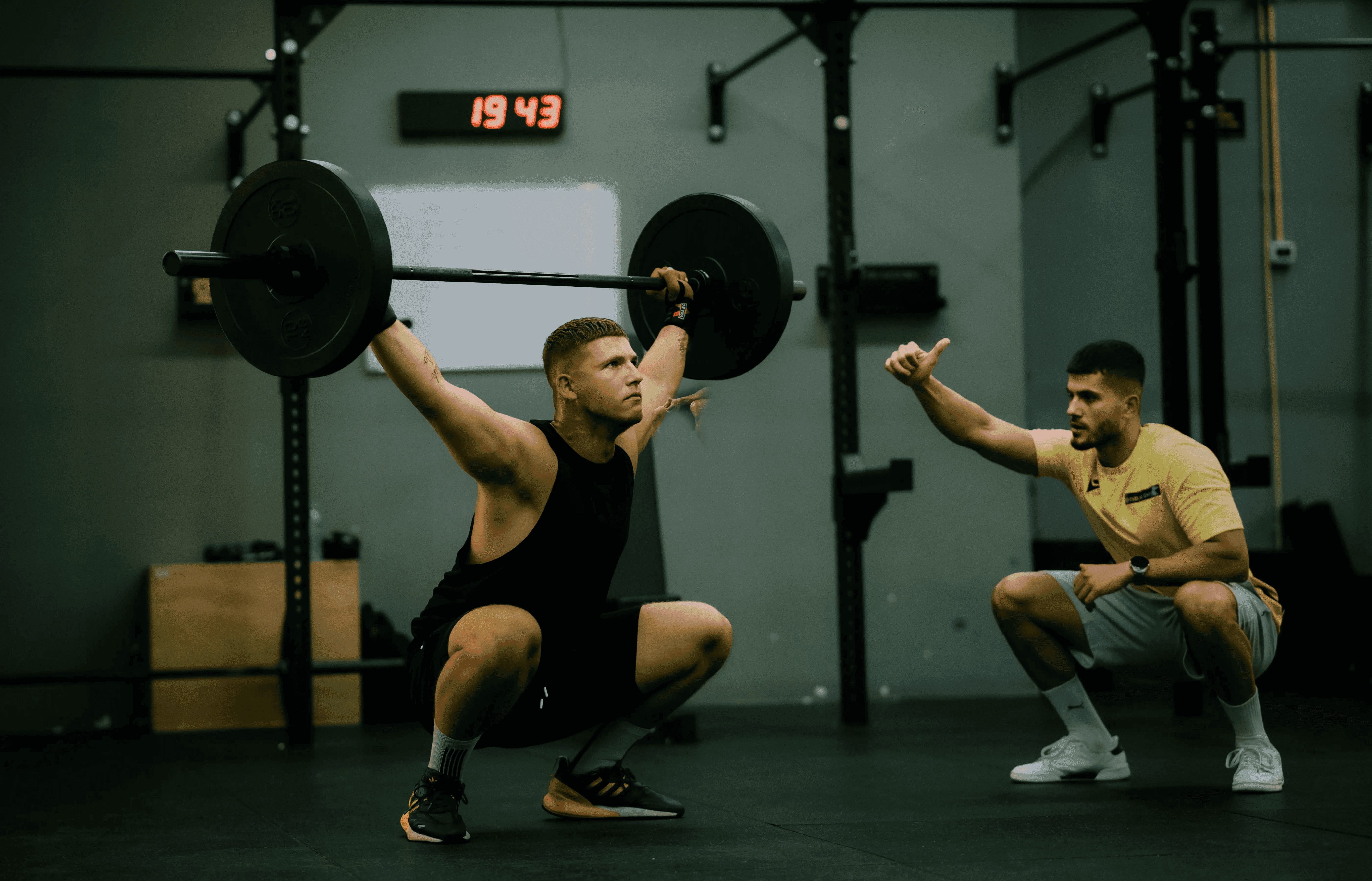 Muscular man in black tank top overhead pressing barbell while coach in yellow shirt squats beside him giving thumbs-up in gym.