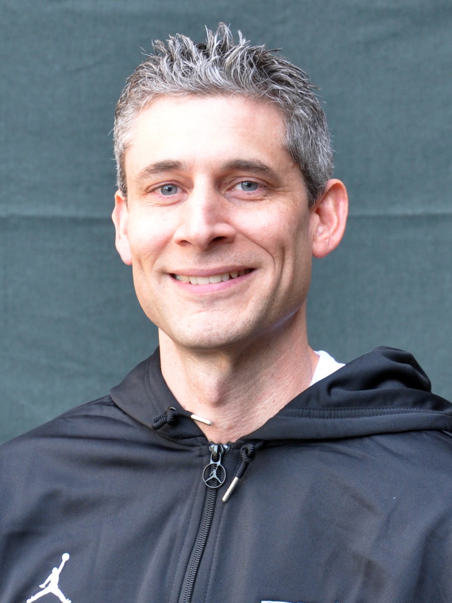 Blonde woman in a black long-sleeve and apron, arms crossed—minimalist studio portrait on a beige background.