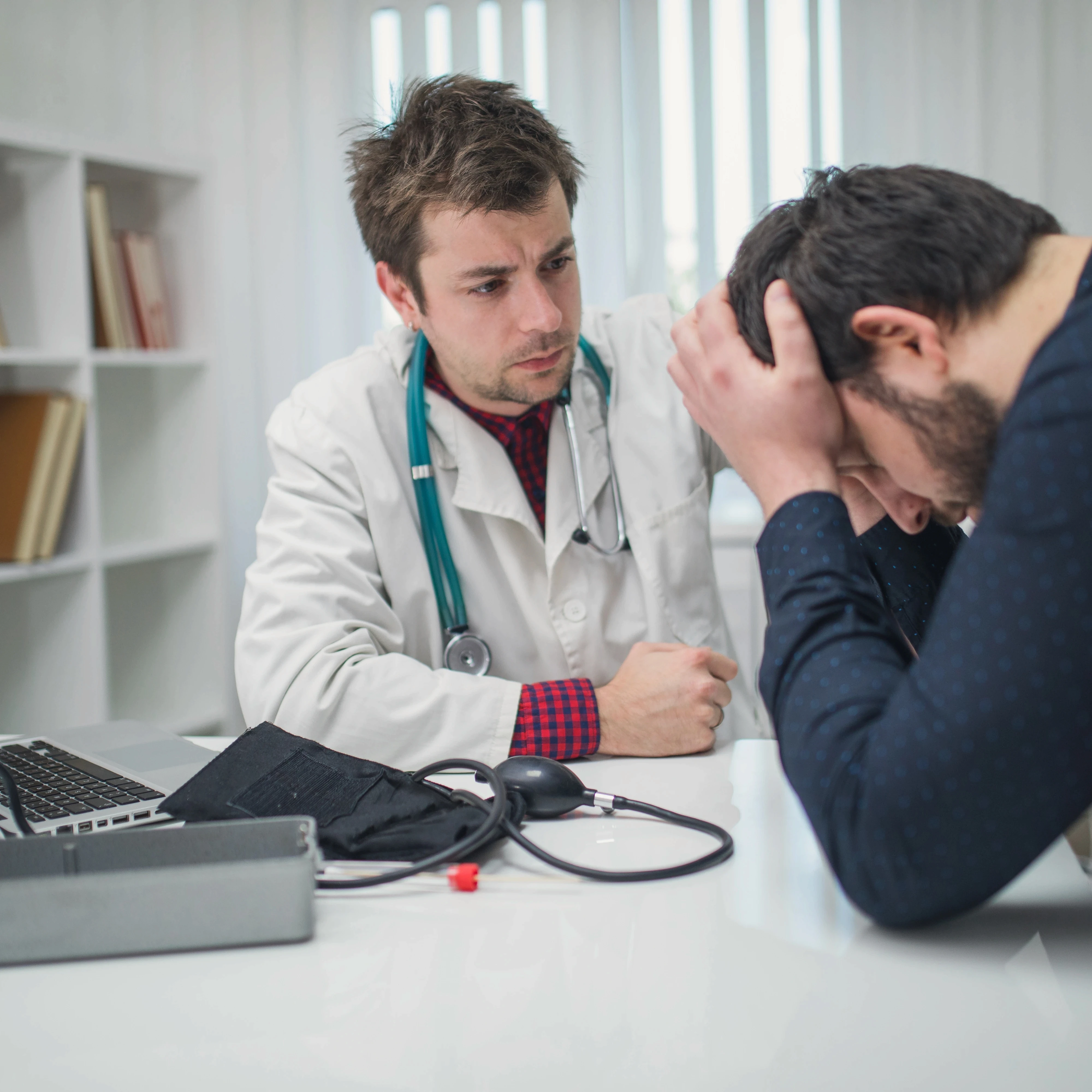 A doctor in a white coat and stethoscope comforts a distressed man with his head in his hands. The setting is a medical office, conveying empathy.