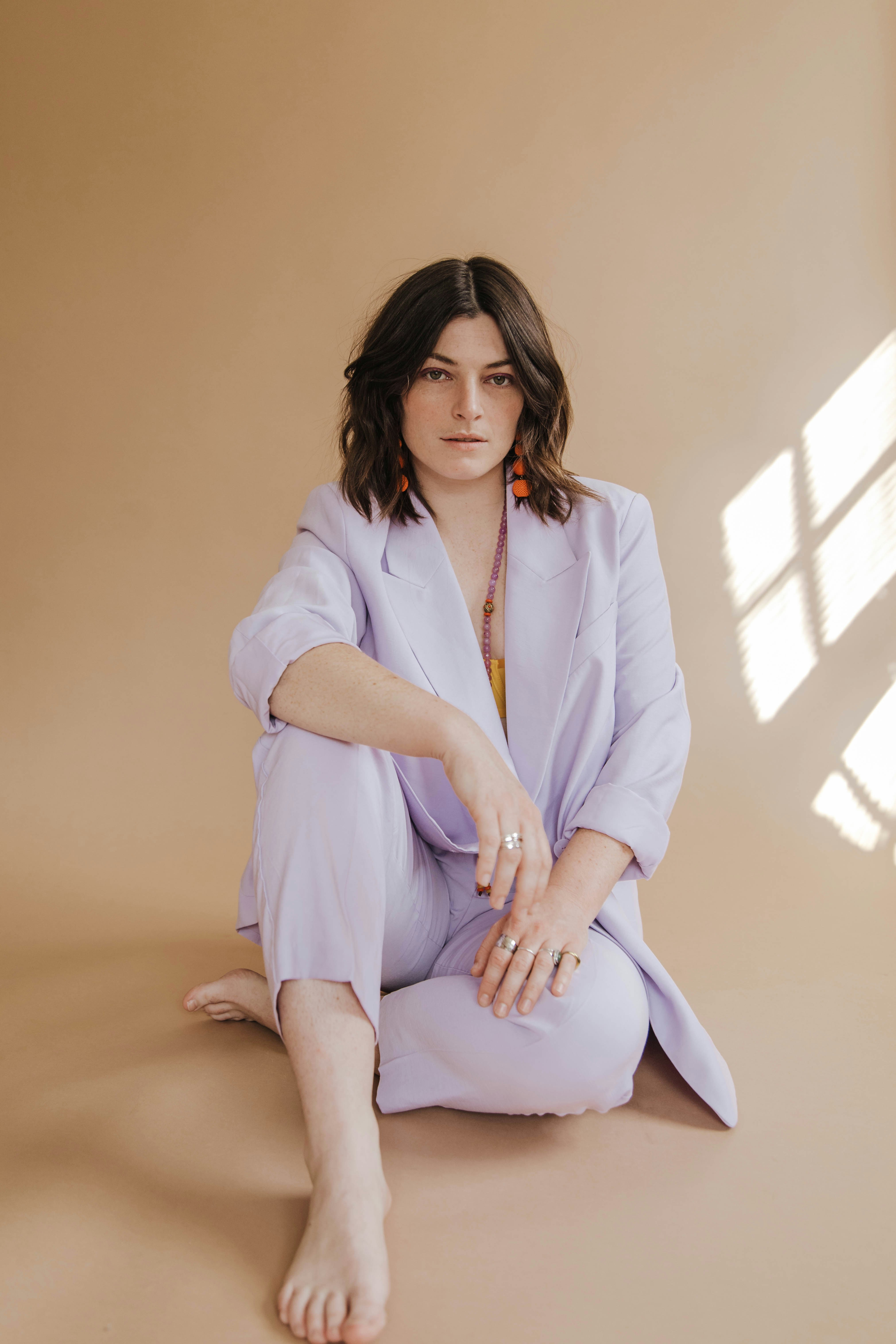 Brunette woman in lilac suit seated on tan backdrop. She wears orange earrings & rings.