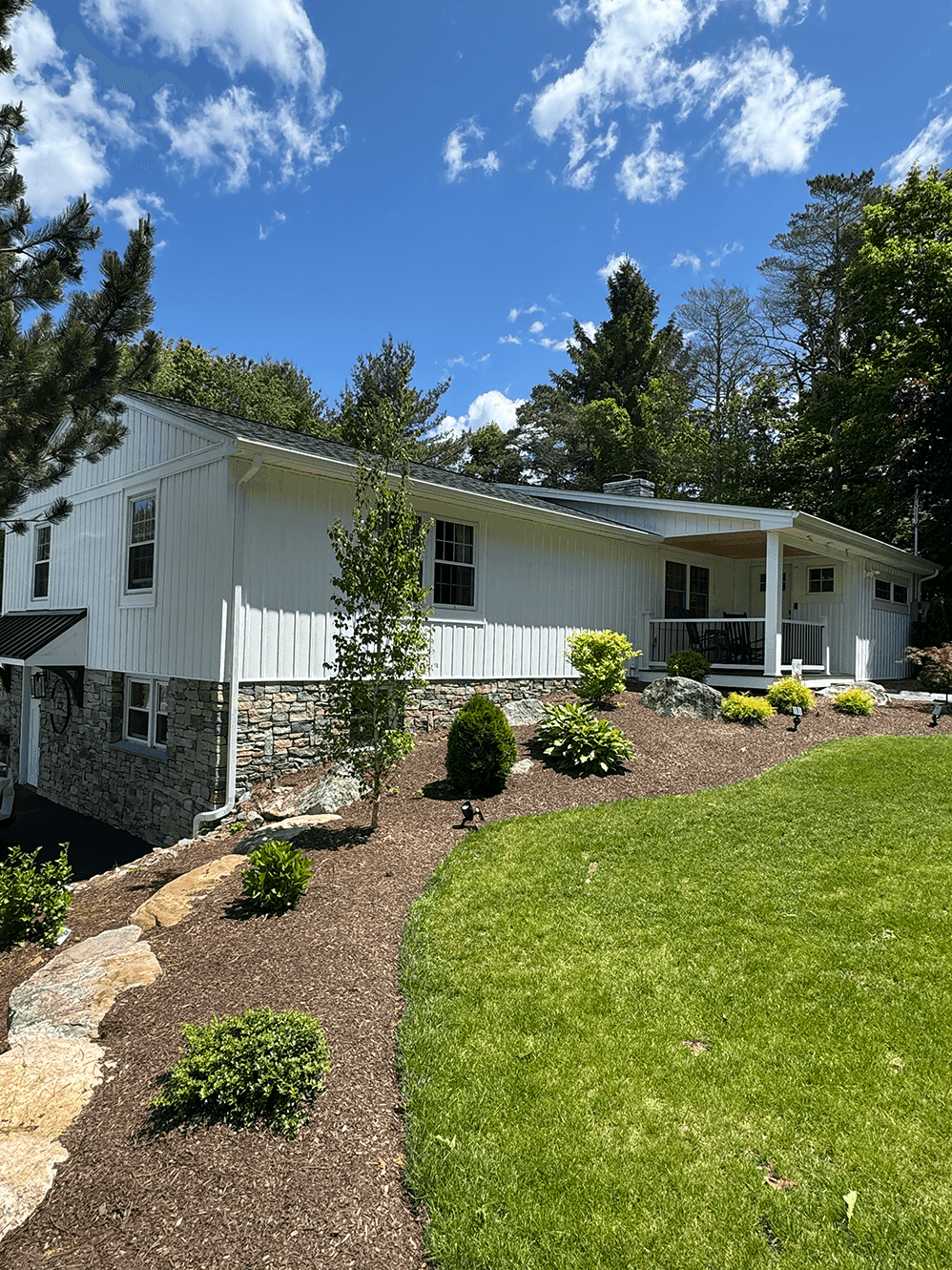 A split-level white home with a stone foundation, surrounded by manicured landscaping and a sloped yard. The front porch includes a seating area, with shrubs and mulch beds enhancing the curb appeal.