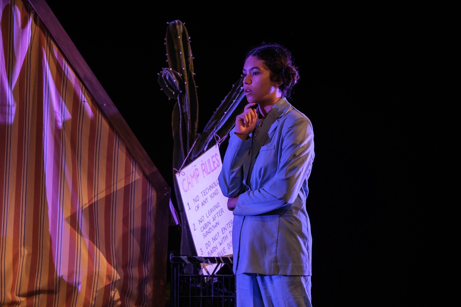A performer in a blue suit stands deep in thought beside a striped tent and a large fake cactus. A handwritten “Camp Rules” sign hangs nearby under purple stage lighting.