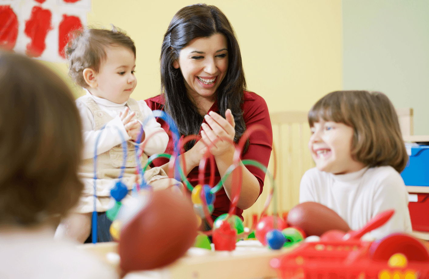 Eine fröhliche Erzieherin spielt mit lachenden Kindern in einer bunten, spielzeugreichen Umgebung, vermutlich in einem Kindergarten oder einer ähnlichen Betreuungseinrichtung.